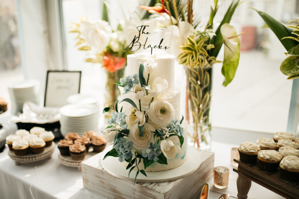 a beautiful wedding cake with sugar flowers and a cake topper featuring the couples name. Surrounding the cake are cupcakes and flowers
