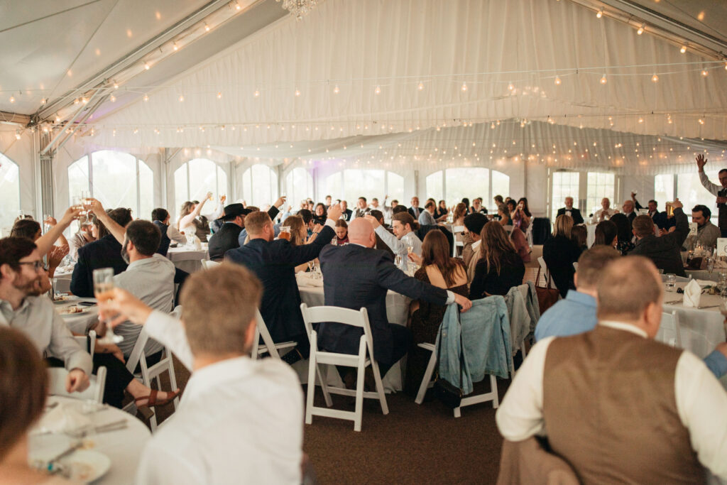 wedding guests inside an event tent with string lights raising their glasses for a toast