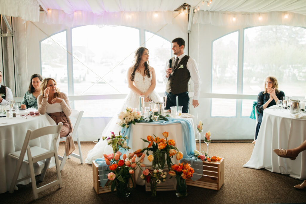 image shows a bride and groom speaking to their guests at the wedding reception from their florally adorned sweetheart table
