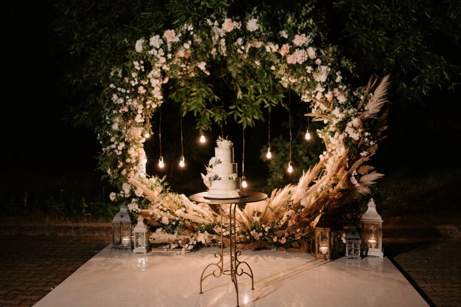 image shows a circular ceremony arch adorned with greenery and lights. A cake stands front and center
