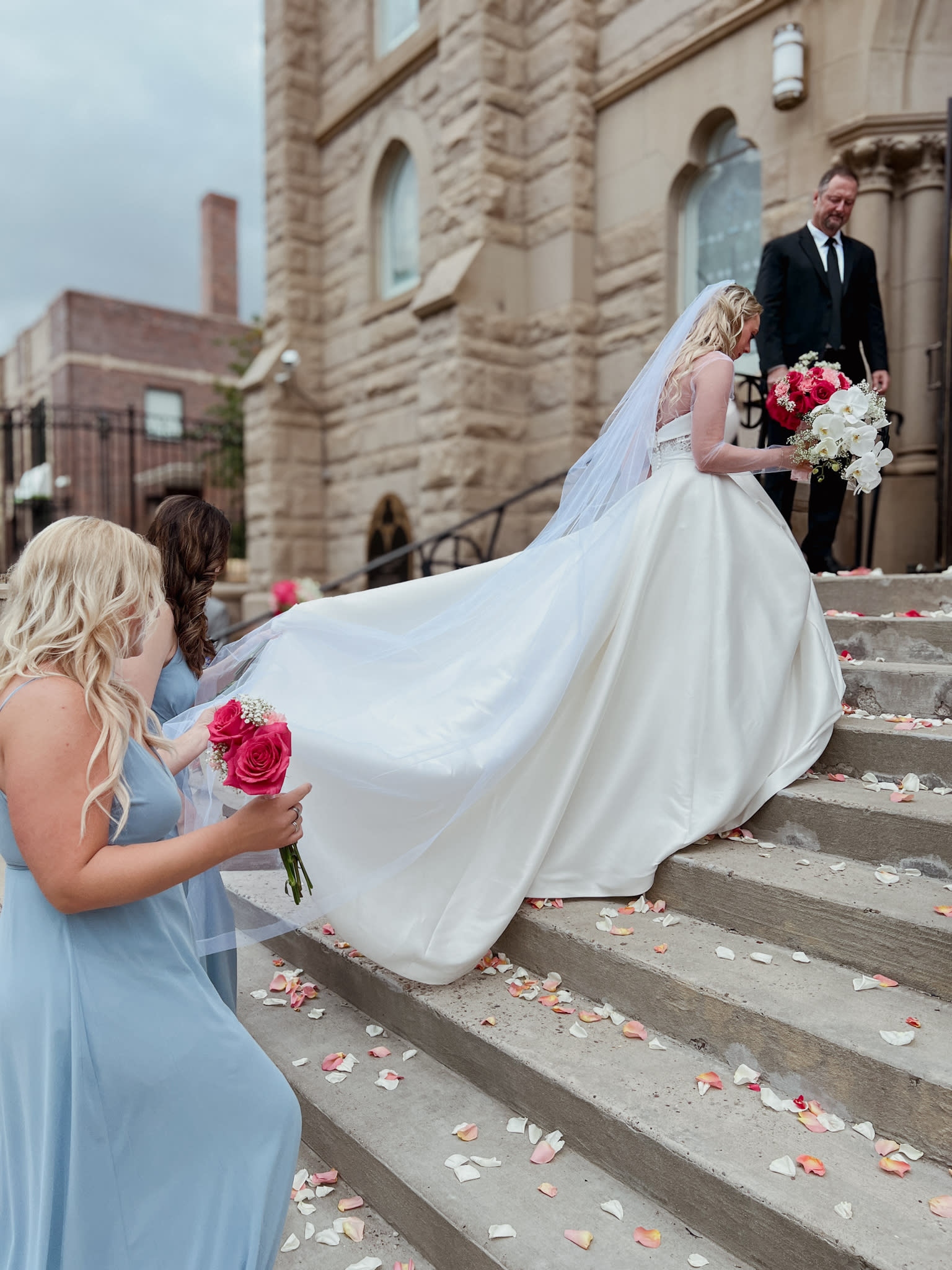 image shows a bride walking up steps with her bridesmaids assisting