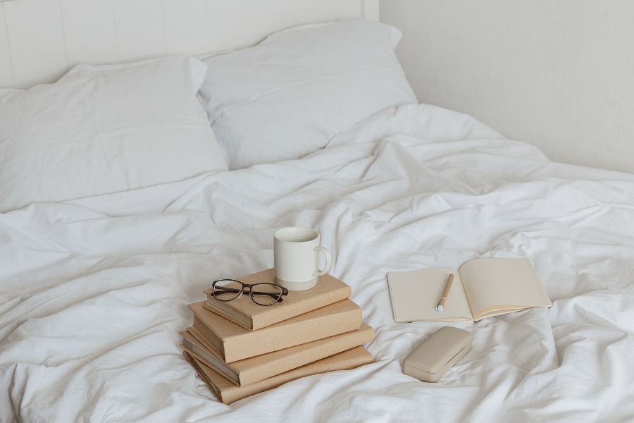 image shows a white bed with cozy bedding, books, glasses, and a mug.