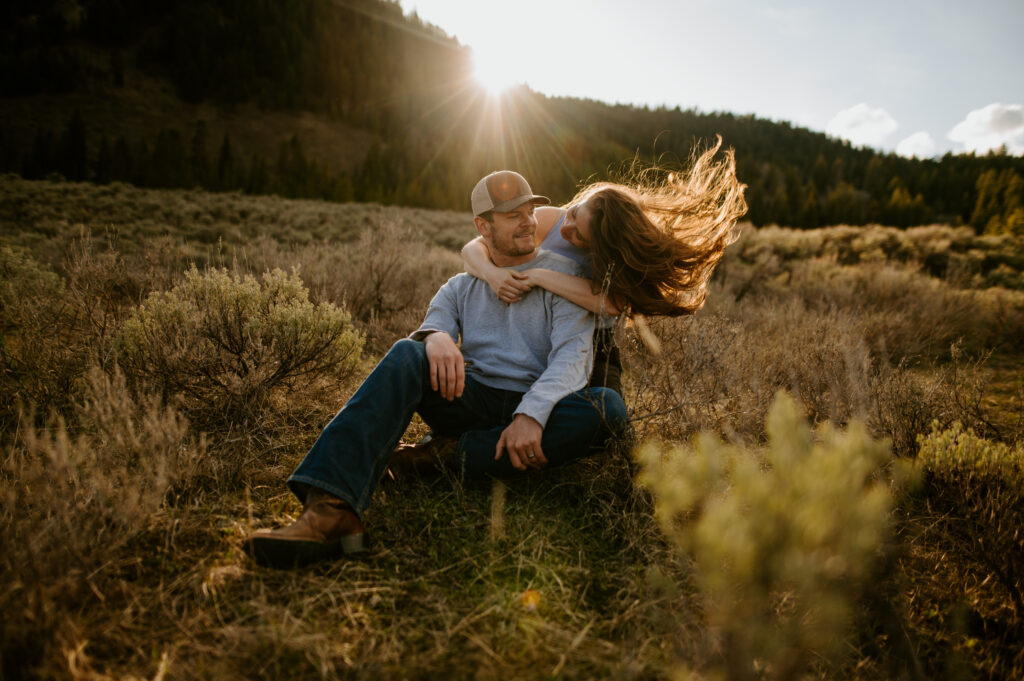 a couple sitting in sage grass posing naturally for a photo. The sun beams behind them while the woman flips her hair.