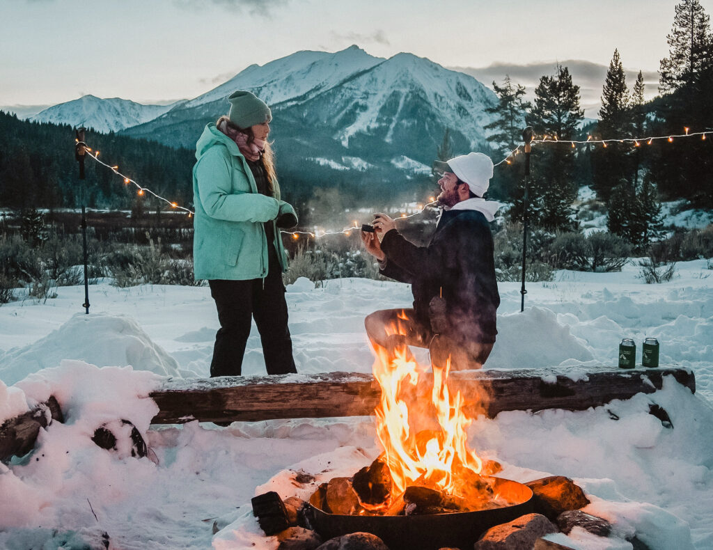 Image shows a man proposing to his partner in front of a fire in the mountains