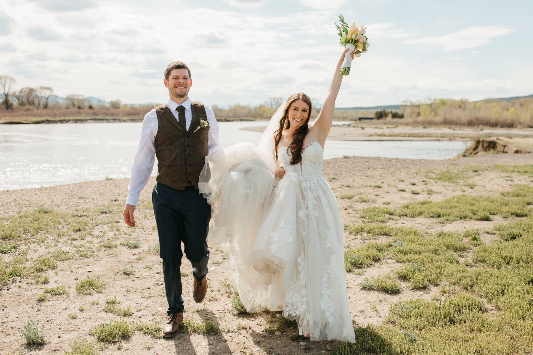 image shows bride and groom walking towards photographer. Bride holds her bouquet up in the air while groom helps bride carry her dress train