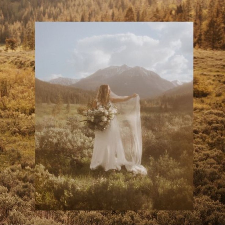 a moody and filmy photo featuring a bride in a bridal gown holding a large bouquet of flowers in one hand while opening her veil in the other. Mountains are the backdrop
