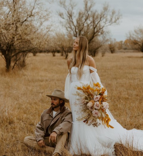 A bride and groom pose for a photo. The groom is sitting on the ground while the bride stands in a dress, holding a large bouquet. 