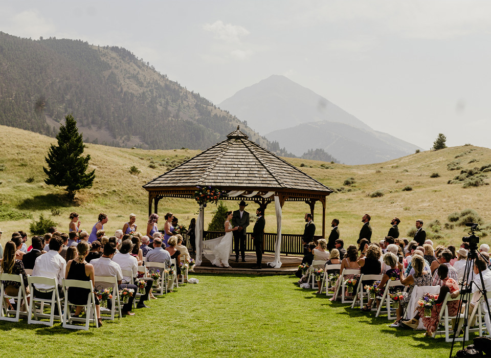 image shows an outdoor wedding under a gazebo with guests looking on. Mountains set the backdrop for this ceremony