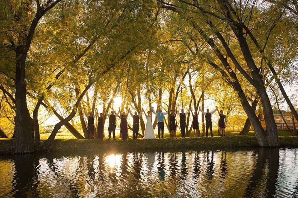 image shows a bridal party holding their hands up in the air with trees surrounding them and a pond showing their reflection. 