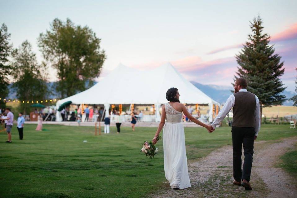 image shows a bride and groom holding hands and walking towards their wedding reception tent where guests await their arrival.