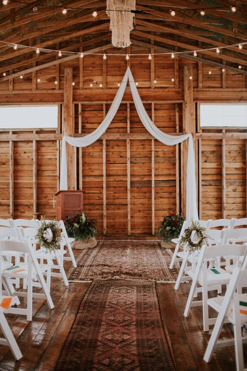 image shows an inside ceremony set up  with rustic barn walls, white chairs and a beautifully draped cloth for the ceremony arch.