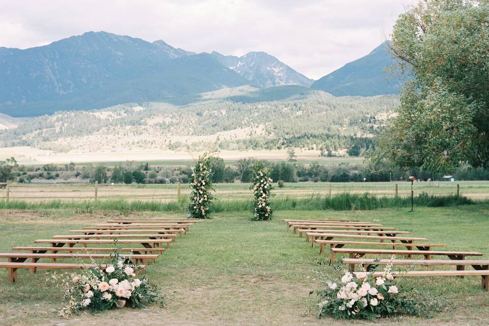 image shows an outdoor wedding ceremony set up with bench seating, floral arrangements set up against a mountain backdrop