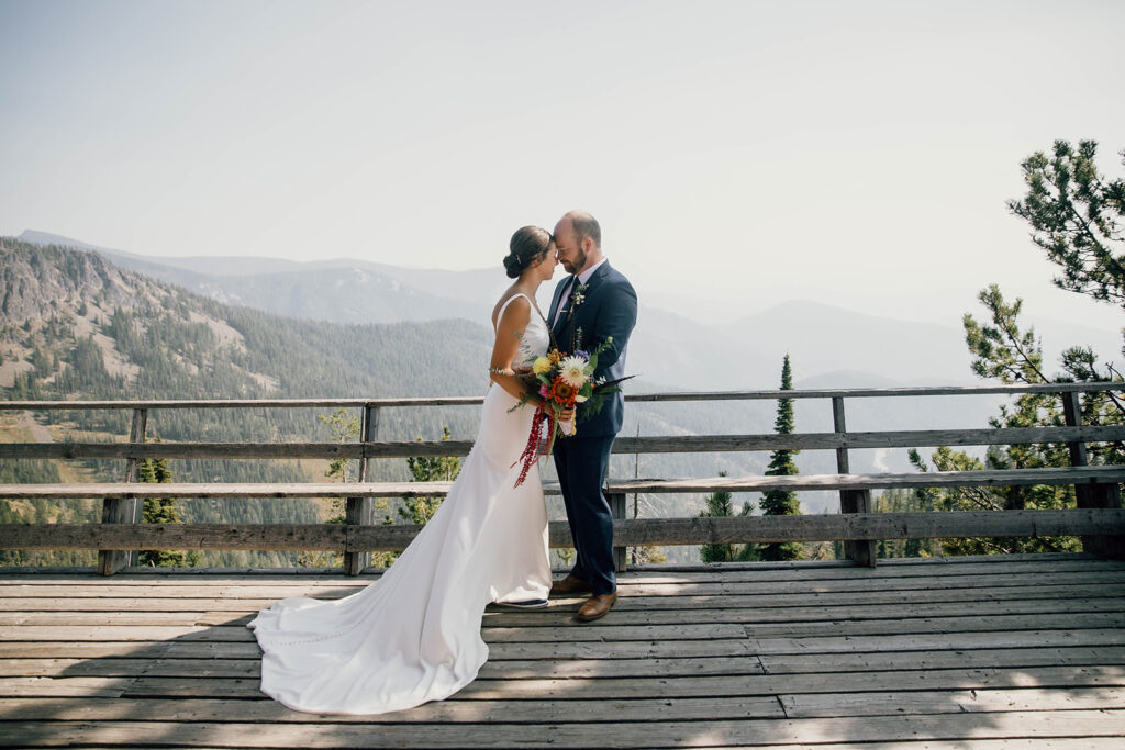 image shows a bride and groom nuzzling heads on deck with a mountain vista