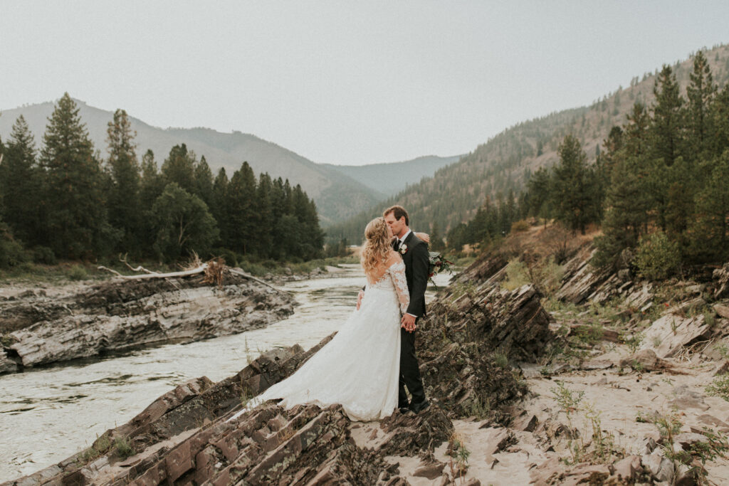 this image shows a bride and groom sharing a kiss alongside a rocky rivers edge
