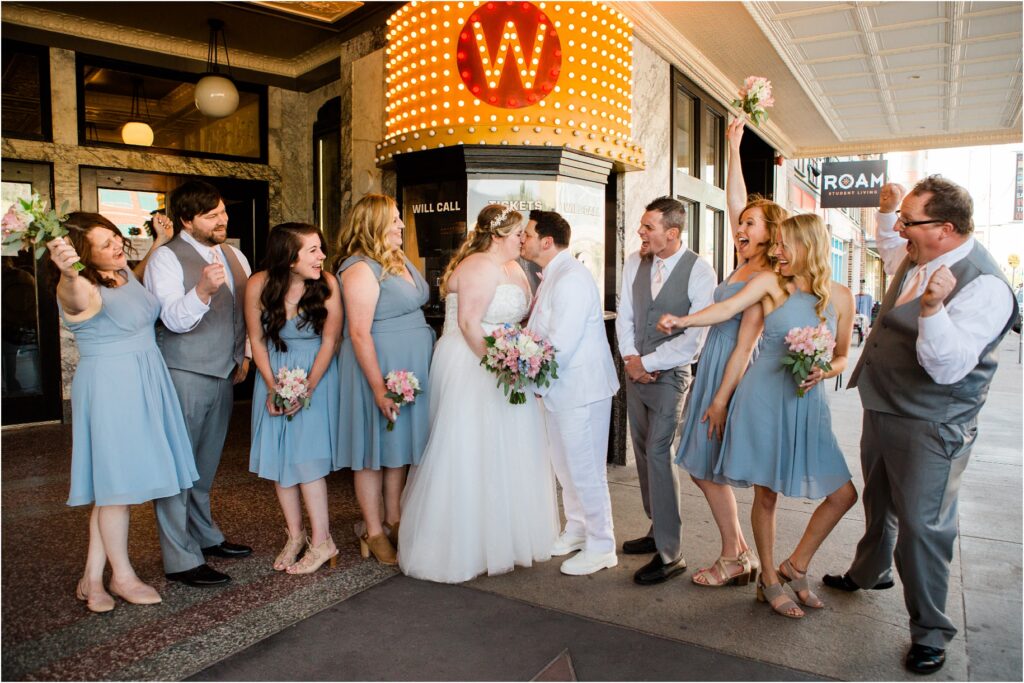 image shows a bride and groom sharing a kiss while their bridal party cheers them on