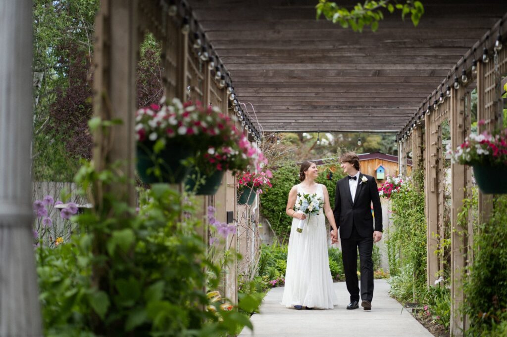 image shows a bride and groom walking hand in hand under a wooden pergola and surrounded by greenery