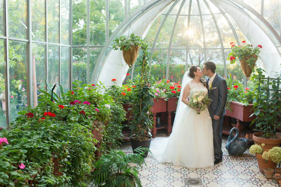 image shows a bride and groom inside a large windowed botanical room surrounded by lush greenery